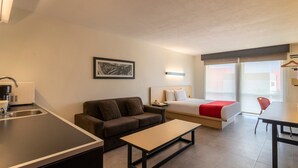 A modern hotel room with a kitchenette, a sofa, a bed with a red accent, a dining table, and a chair. - City Express by Marriott Chetumal (Chetumal)
