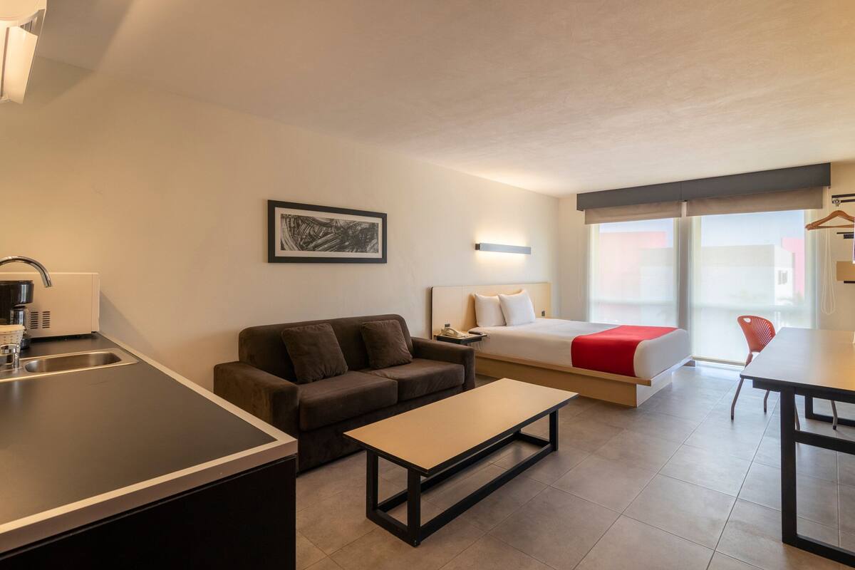 Una habitación de hotel moderna con kitchenette, un sofá, una cama con detalles en rojo, una mesa de comedor y una silla.