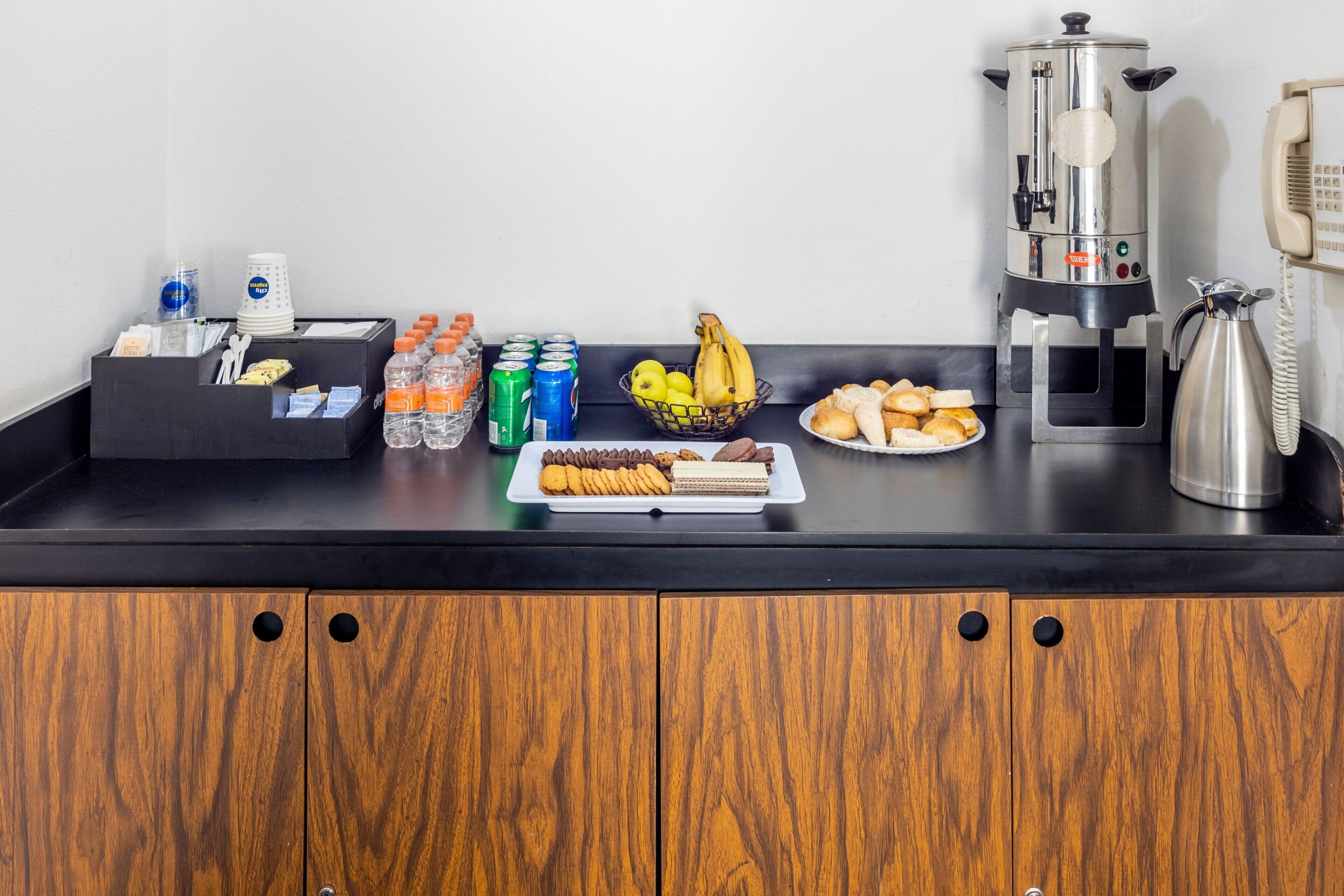 A hotel reception area with a black counter, wooden cabinets, a coffee machine, a phone, and various food items.