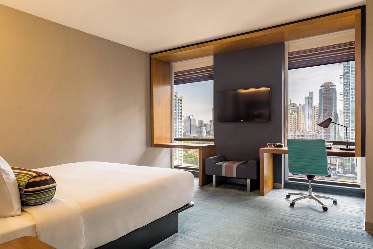 A hotel room with a bed, a desk with a chair, a TV mounted on the wall, and large windows offering a city view.