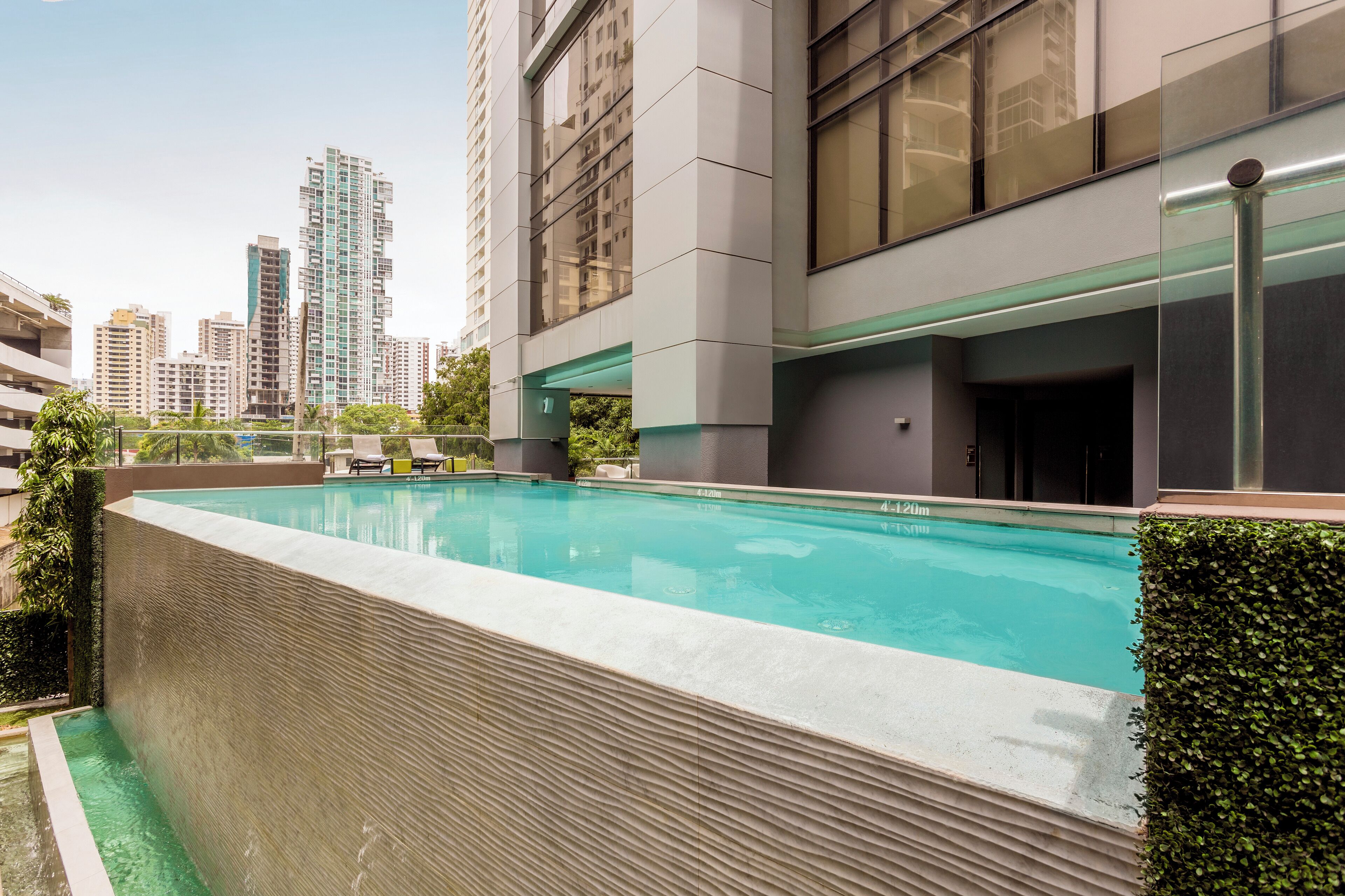 Un edificio moderno con piscina en la azotea, rodeado de vegetación y edificios altos al fondo.