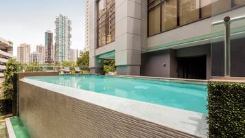Un edificio moderno con piscina en la azotea, rodeado de vegetación y edificios altos al fondo.