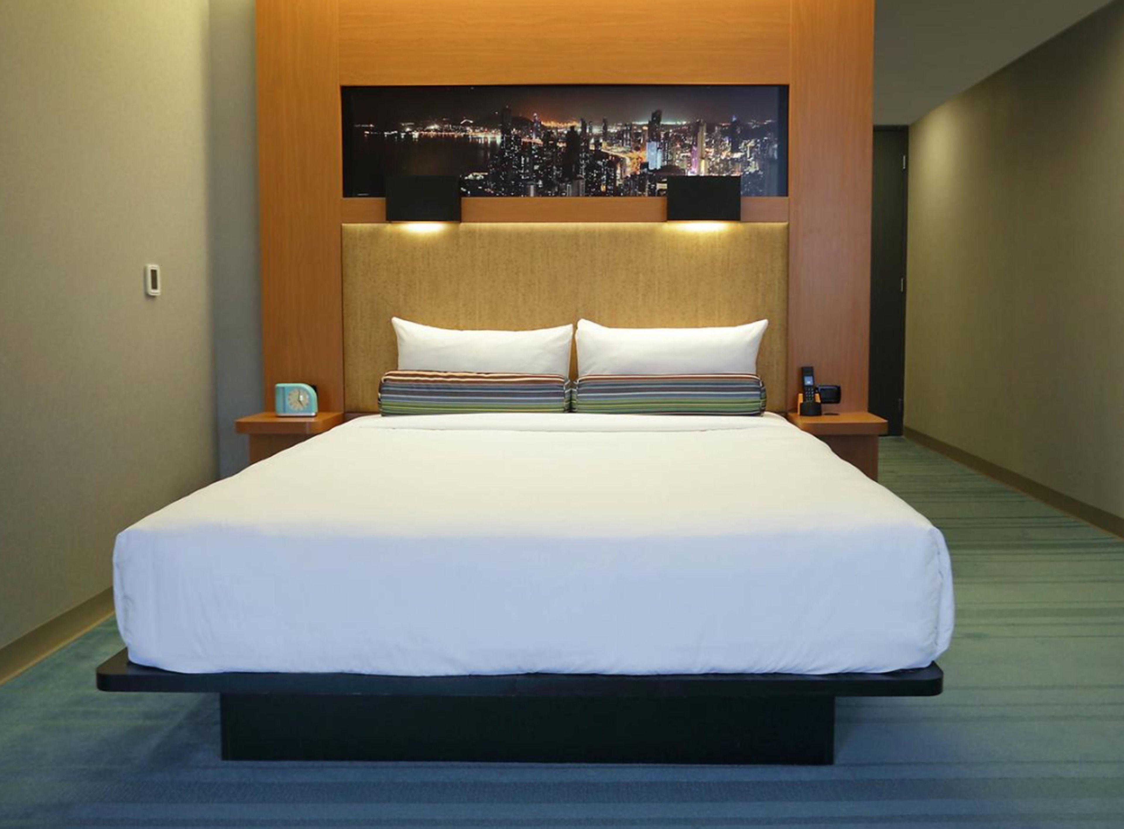 Habitación de hotel con una cama grande, dos almohadas, una mesita de noche con un reloj y una obra de arte urbana sobre la cama.