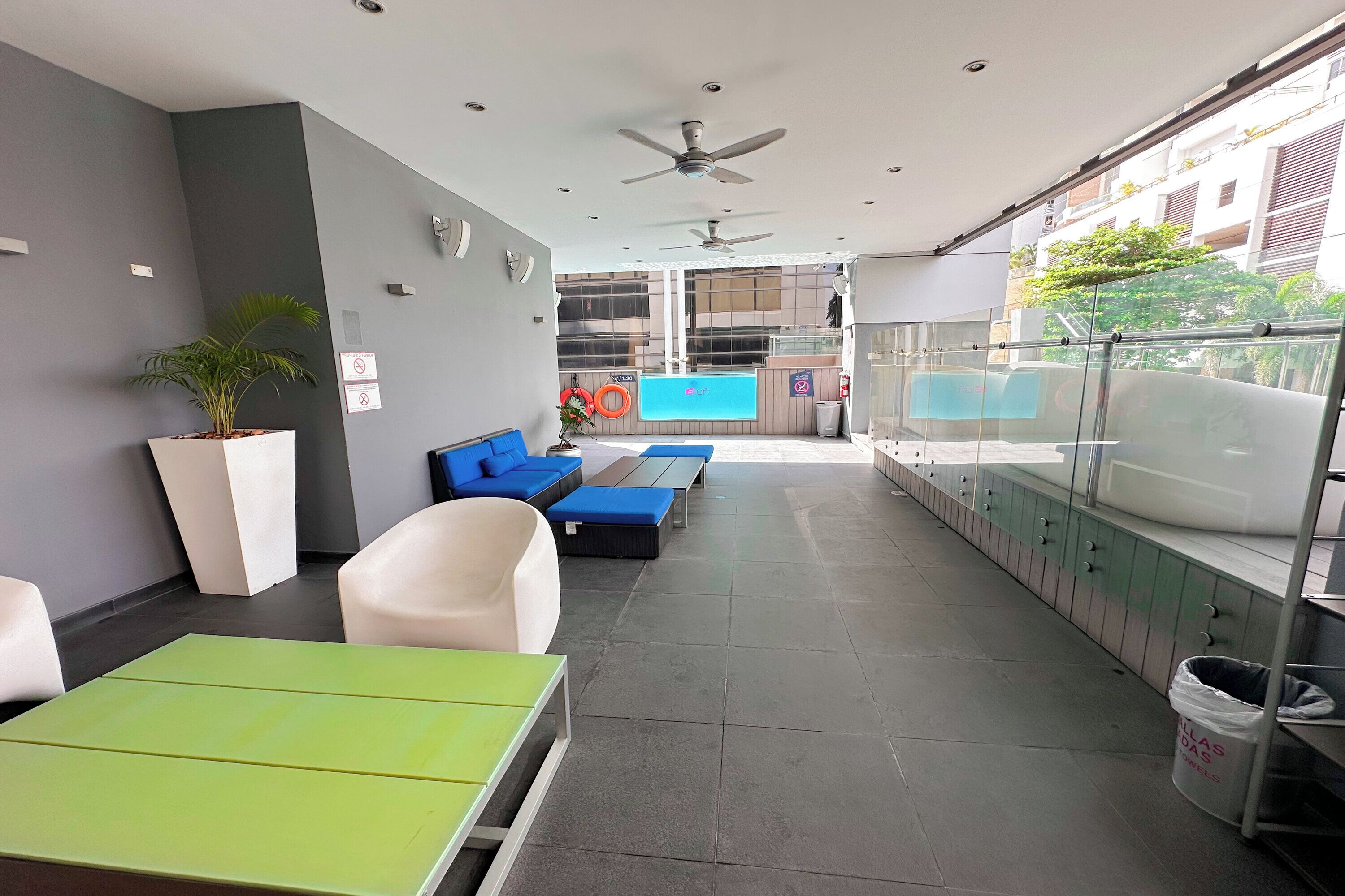 Un área de estar interior moderna con ventilador de techo, una planta en maceta y un amplio ventanal con vistas a un edificio.