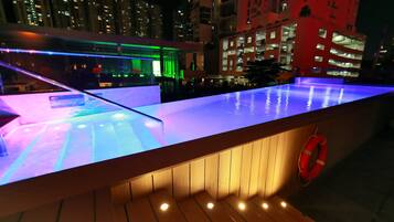 A rooftop pool area with a glass barrier and wooden steps, surrounded by high-rise buildings.
