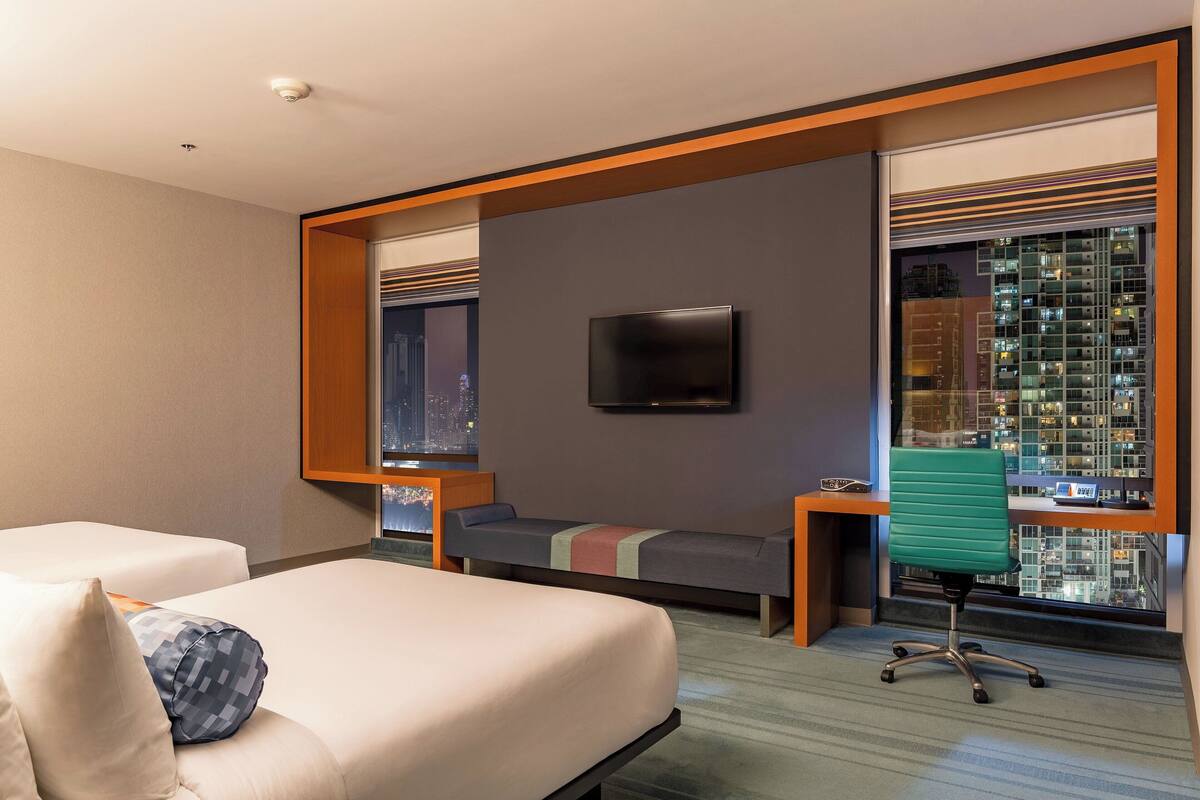 A modern hotel room with a large bed, a desk, a chair, a TV, and a city view.