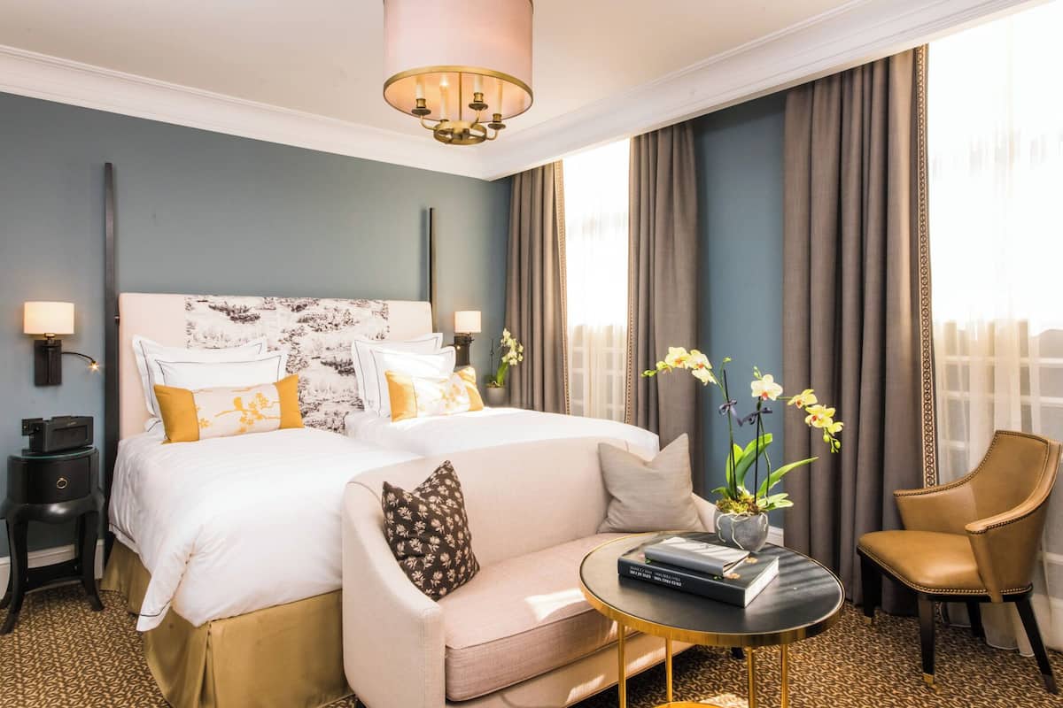 Ein Hotelzimmer mit einem großen Bett, einer Couch, einem kleinen runden Tisch mit Büchern, einem Sessel und einer Zimmerpflanze.
