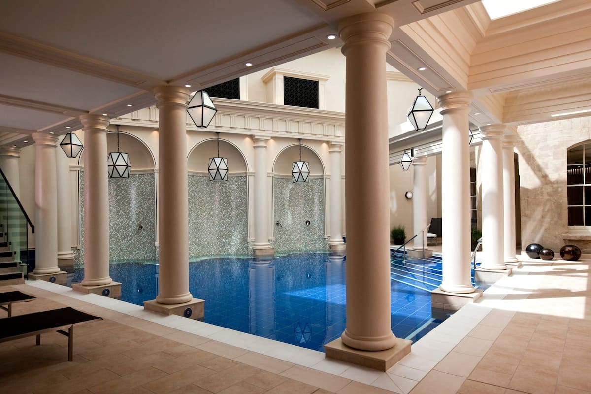 Ein geräumiger, überdachter Pool mit klarem, blauem Wasser, umgeben von weißen Säulen und Bögen, sowie eine Treppe, die zu einer erhöhten Ebene führt.