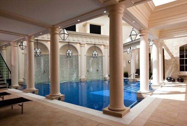 A spacious indoor swimming pool with clear blue water, surrounded by white columns and arches, and a staircase leading to an upper level.