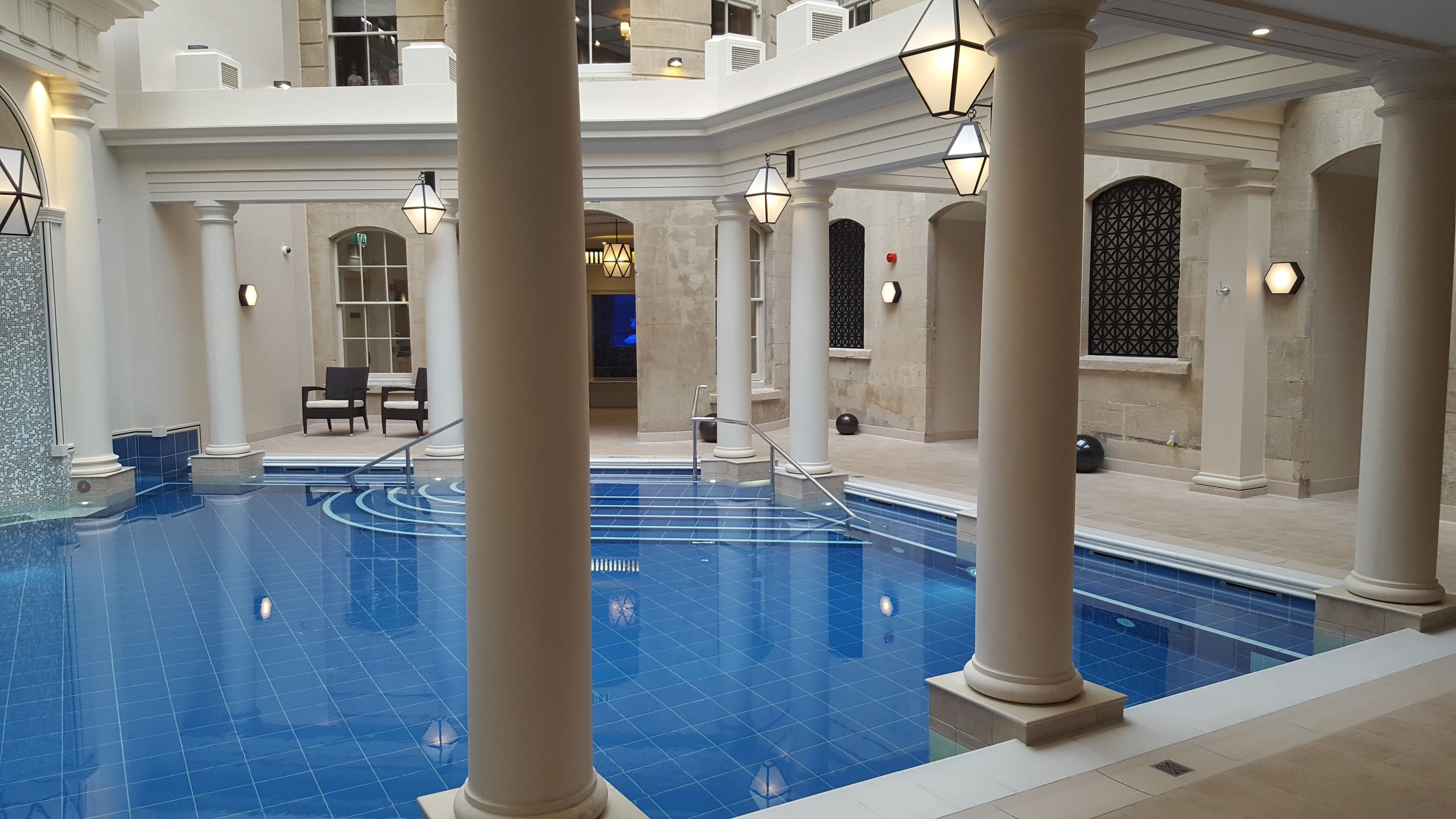 An indoor swimming pool with a tiled floor, surrounded by white columns and arches, and equipped with seating and lighting fixtures.