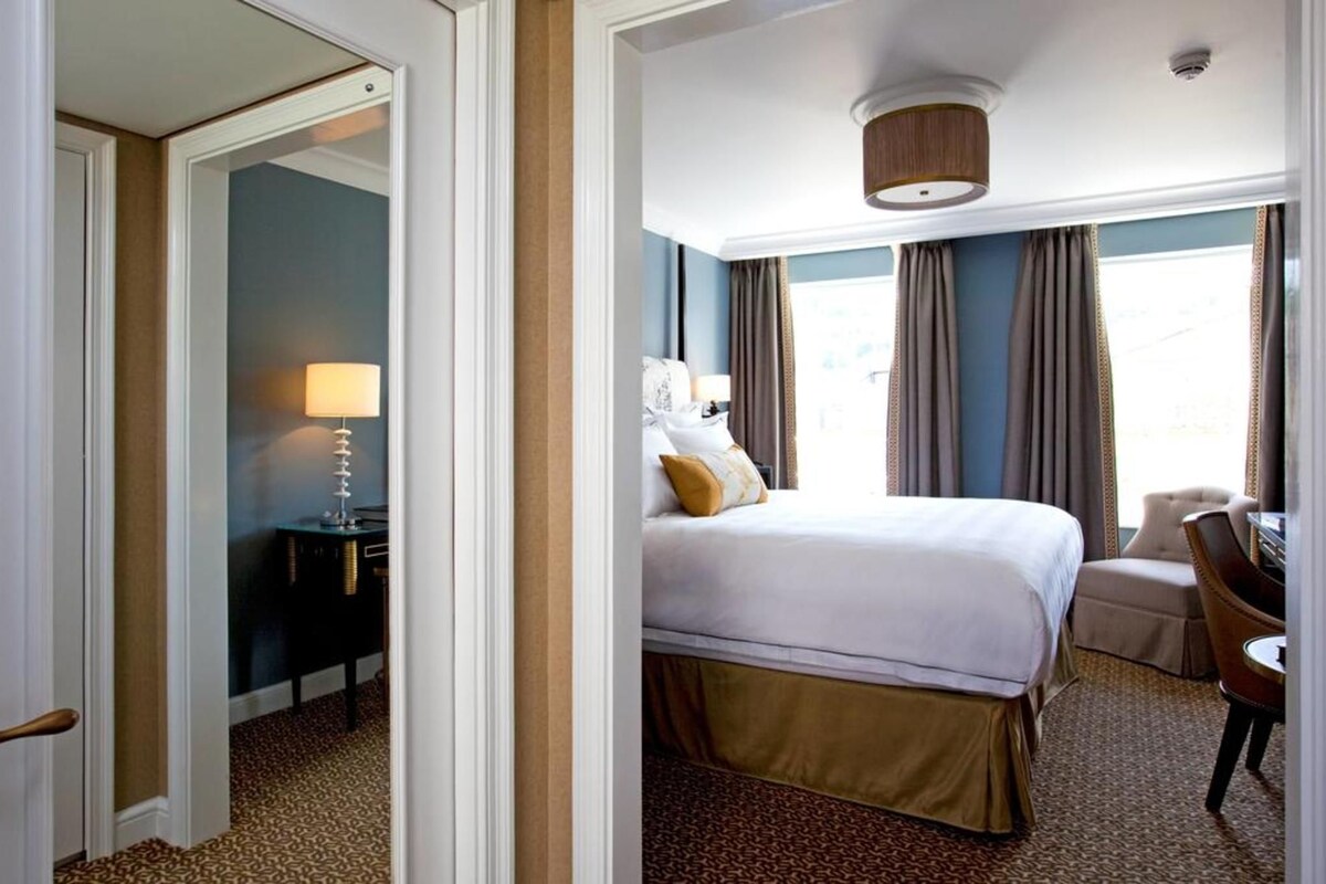 A hotel room with a large bed, a desk with a lamp, a chair, and a view of the outdoors.