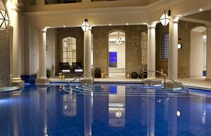 A large indoor swimming pool with a tiled floor, surrounded by columns and arches, and a seating area with chairs and tables.