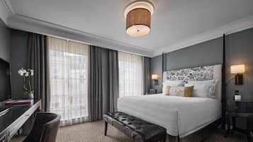 A hotel room with a large bed, a desk with a chair, a bench, and a large window with curtains.