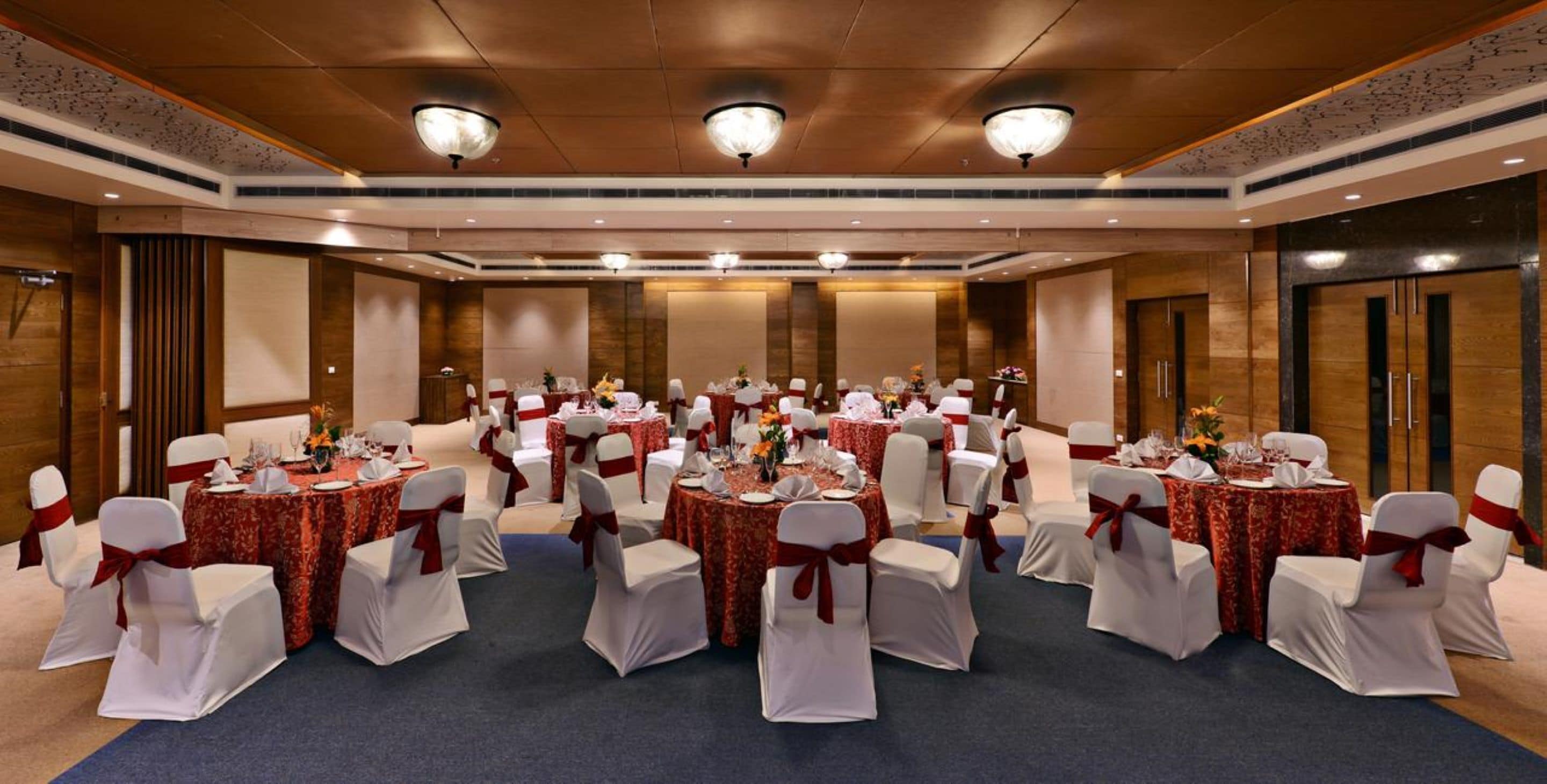 A banquet hall with round tables, white chairs with red sashes, and a blue carpet.