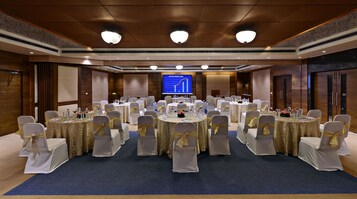 Uma sala de conferências com várias mesas redondas, cada uma com cadeiras brancas e fitas douradas, preparada para uma reunião ou evento.