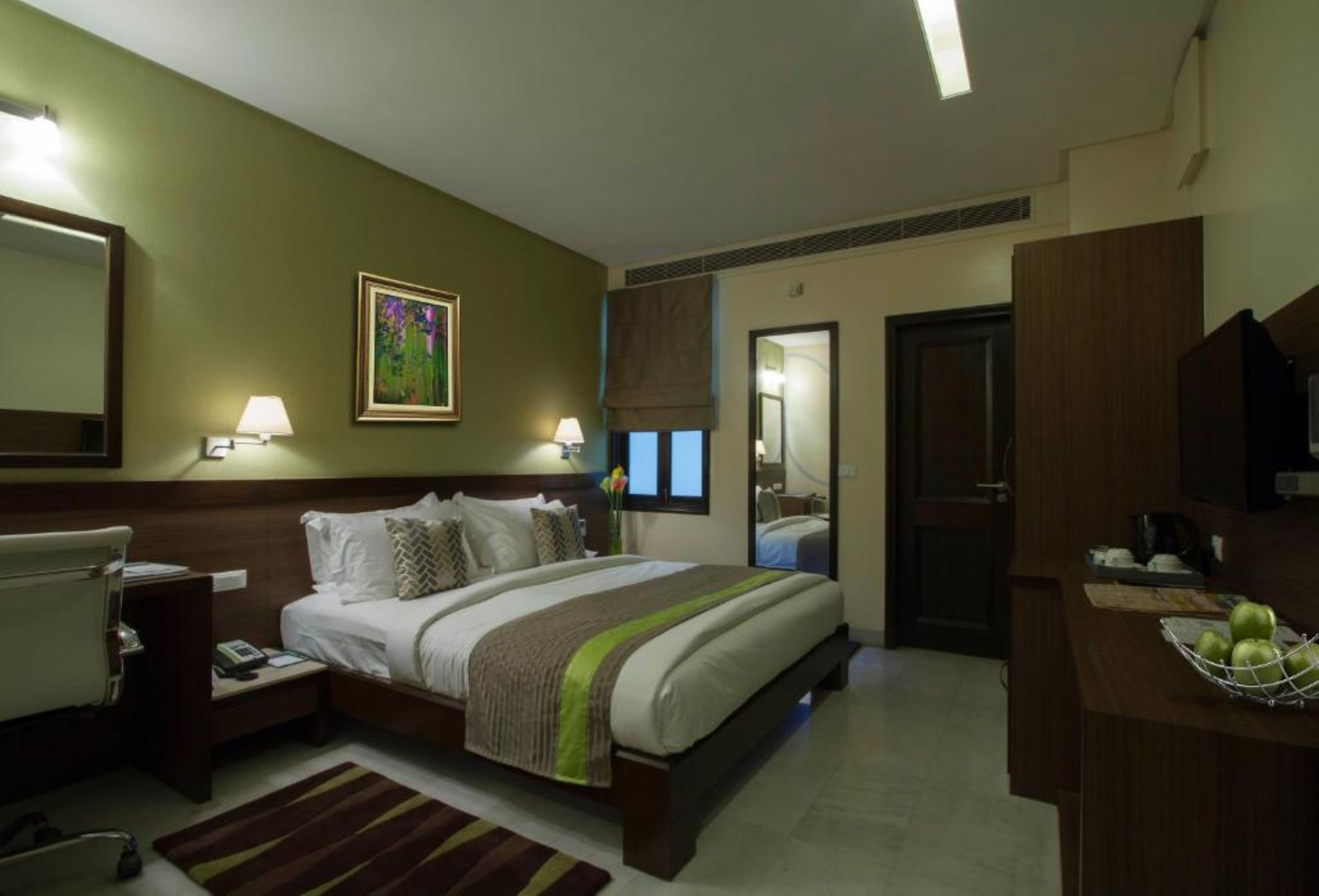 A hotel room with a bed, bedside tables, a desk with a chair, a mirror, and a window with curtains.