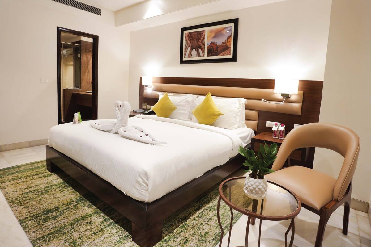 A hotel room with a bed, a chair, a small table with a plant, and a painting on the wall.