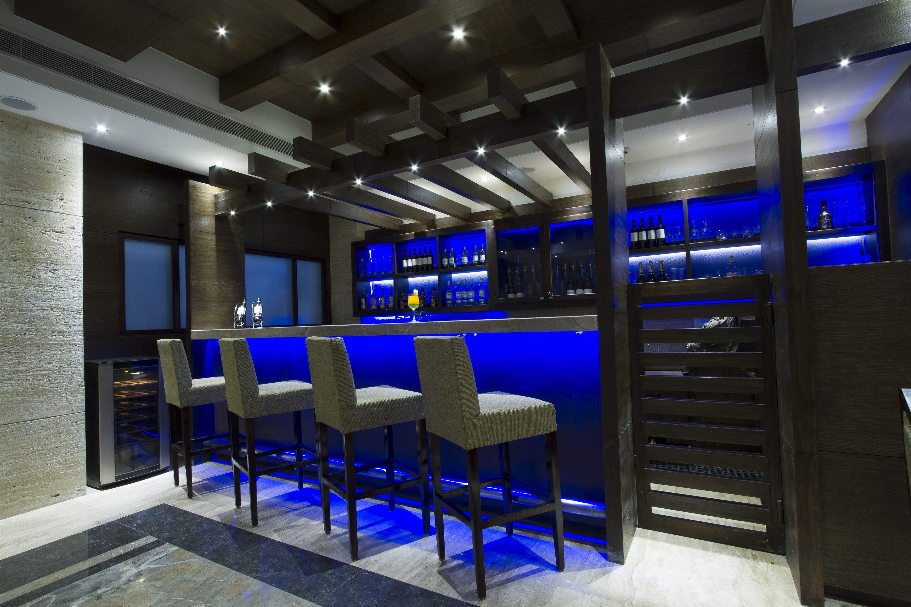 A modern bar area with a dark wood bar counter, blue lighting, and three bar stools.