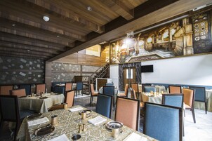 Restaurante com vigas de madeira no teto, paredes de pedra e uma escadaria que leva a um andar superior. Mesas montadas com pratos, copos e talheres.
