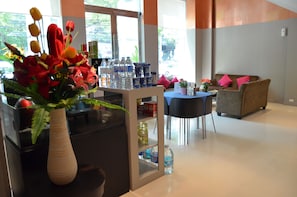 A modern living room with a sofa, a dining table, and a kitchen area with a vase of flowers and water bottles. - Gm Complex (Bangkok)