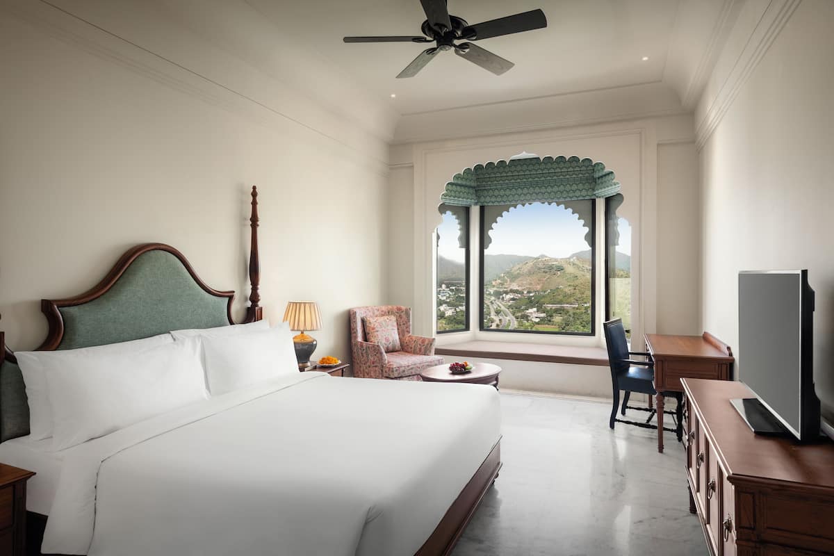 A hotel room with a large bed, a desk, a chair, a television, and a view of the mountains through a window.