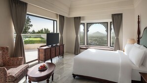 Habitación de hotel con una cama grande, un televisor, una mesa de comedor con fruta y vistas a la ciudad y las montañas.