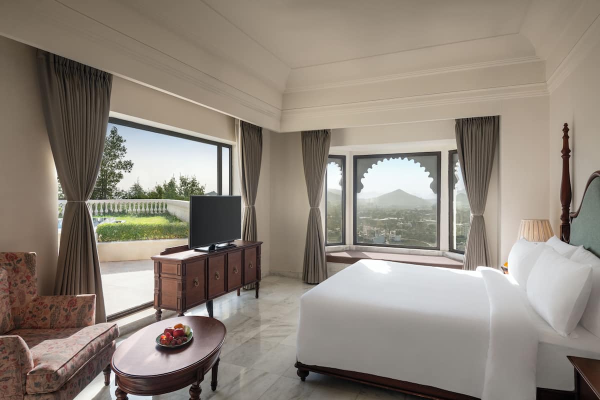 A hotel room with a large bed, a television, a dining table with fruit, and a view of a city and mountains.