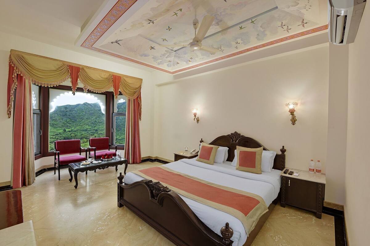 A hotel room with a large bed, two chairs, a table, and a view of mountains through the window.