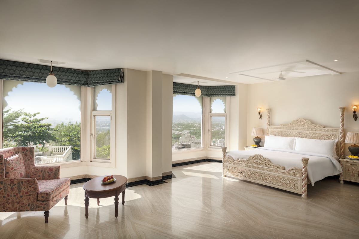 A spacious bedroom with a large bed, a sitting area with a chair and table, and large windows offering a view of the outdoors.