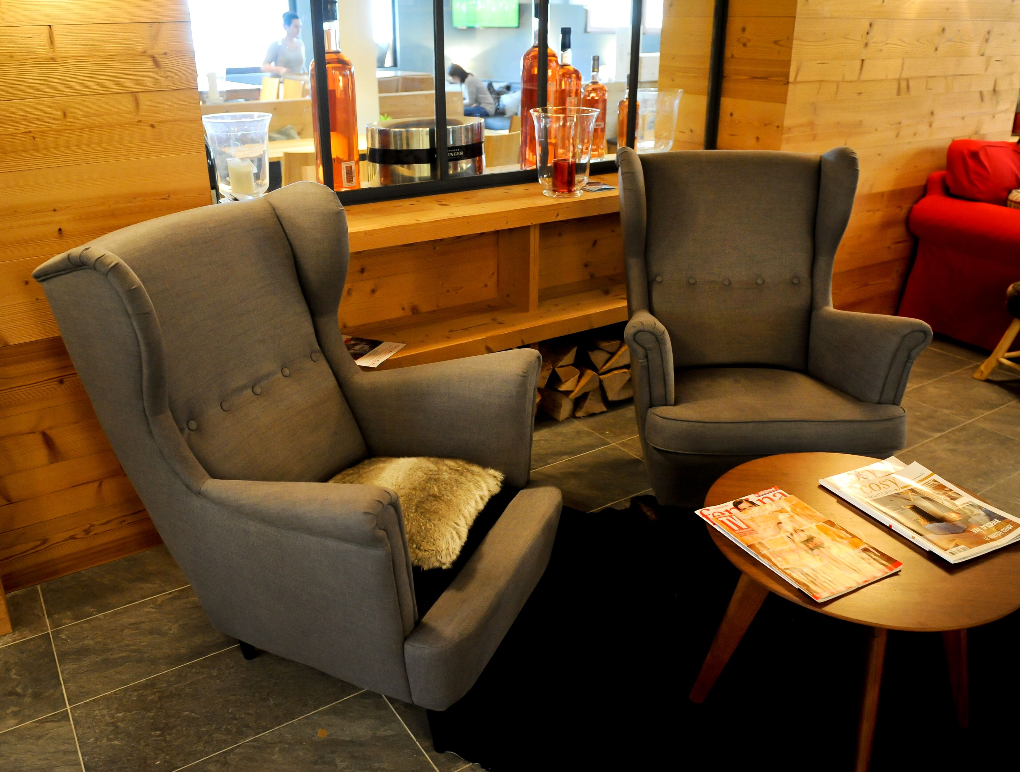 Deux fauteuils gris, une petite table placée entre eux, un banc en bois avec des bouteilles, et une chaise rouge à l’arrière-plan.