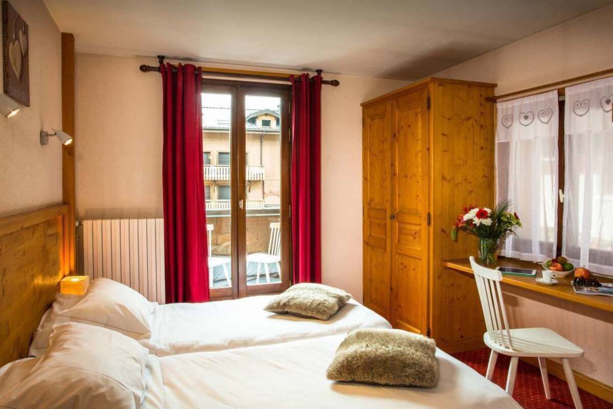 Une chambre d’hôtel avec deux lits, une armoire en bois, un bureau avec une chaise et une fenêtre avec des rideaux rouges.