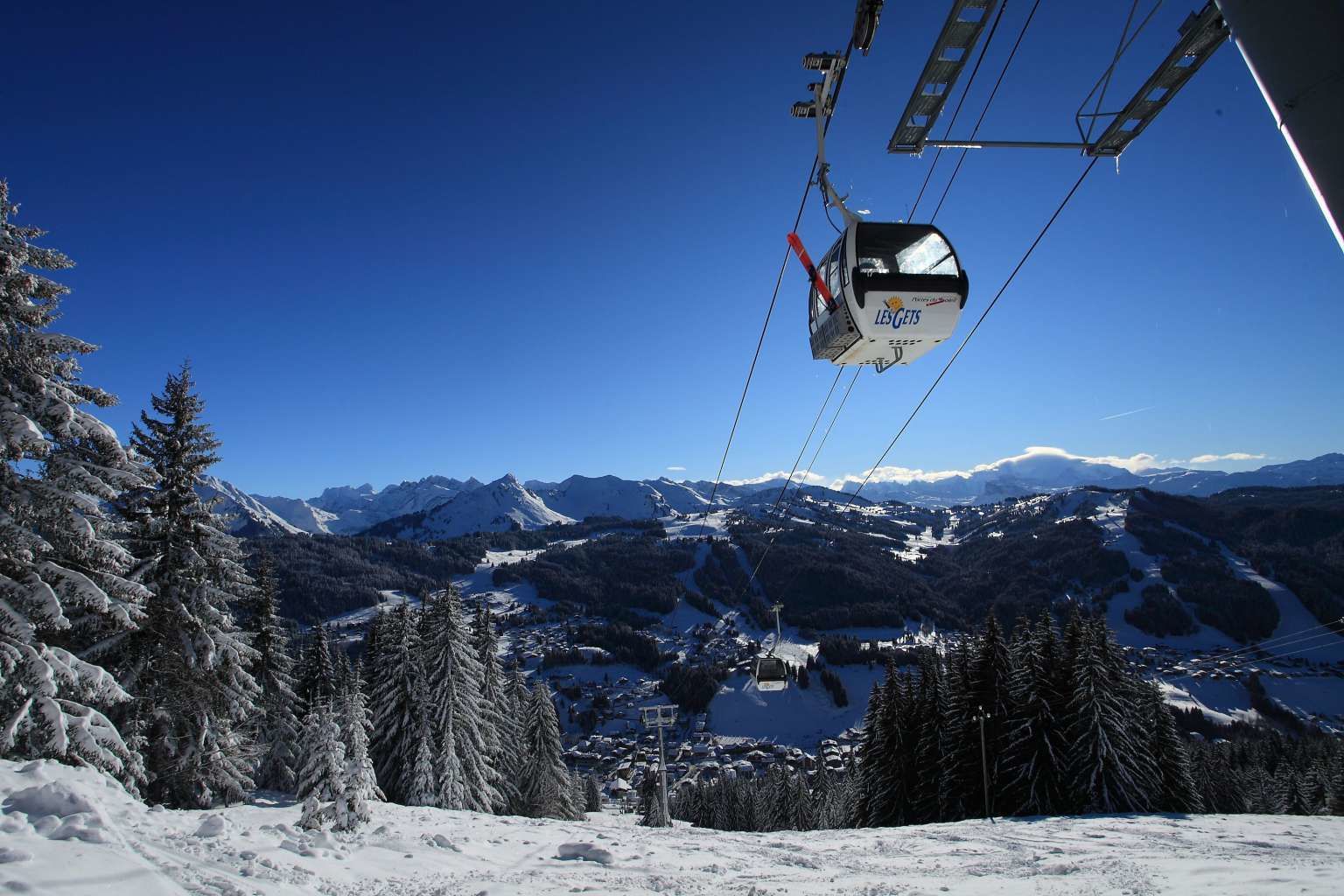 A cable car with 'LESGETS' branding, suspended over a snowy mountain landscape with dense forests and distant peaks.