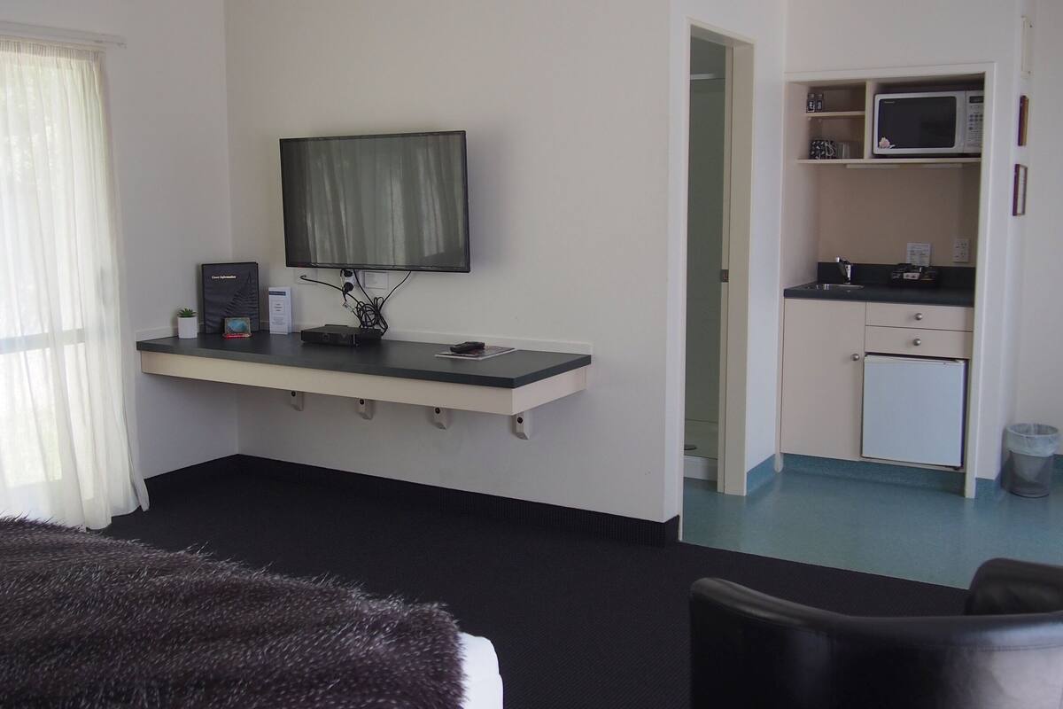A hotel room with a bed, a desk with a computer, a TV, a small kitchenette, and a bathroom.