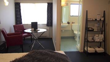 A hotel room with a bed, a red chair, a small table with a plant, a window with curtains, a bathroom with a sink, toilet, and a shelf with towels and toiletries.