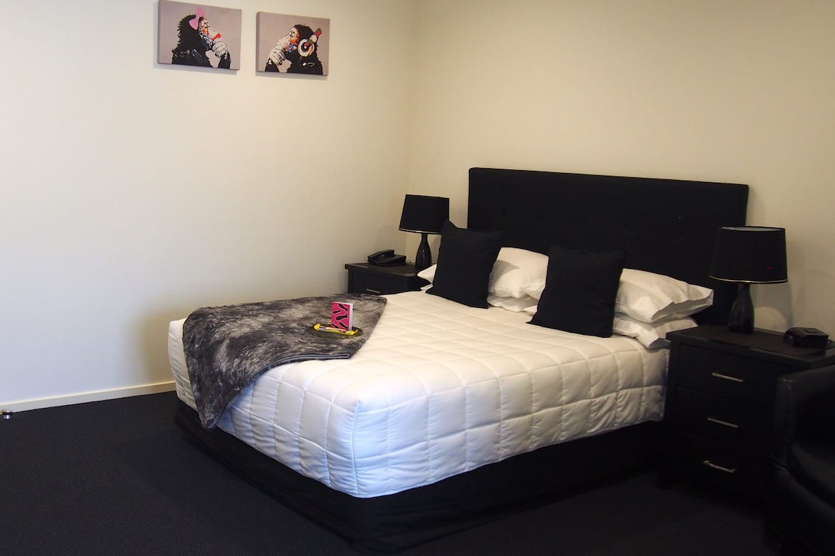 A neatly made bed with a white duvet, black pillows, and a grey throw blanket. There are two bedside tables with lamps and a small nightstand with a phone. The room has a simple, clean design with a dark carpet and white walls.