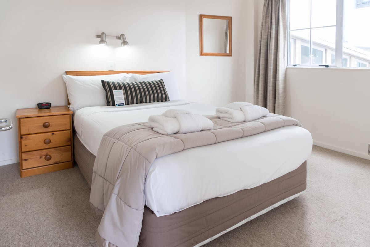 A neatly made bed with a wooden headboard, a bedside table with drawers, a wall-mounted lamp, a window with curtains, and a mirror on the wall.