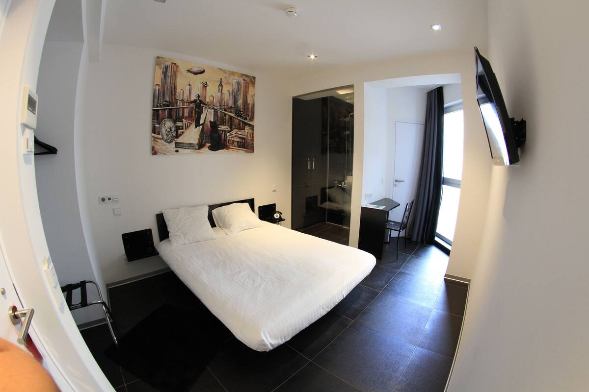 A modern hotel room with a large bed, a painting on the wall, a TV mounted on the wall, and a bathroom visible through an open door.