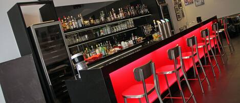 A bar area with a red bar counter, multiple shelves of bottles, and several bar stools.