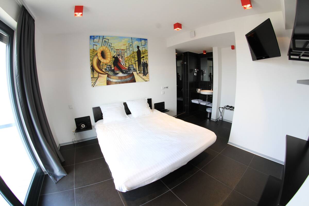 A modern hotel room with a large bed, a painting on the wall, a TV mounted on the wall, and a bathroom visible through an open door.