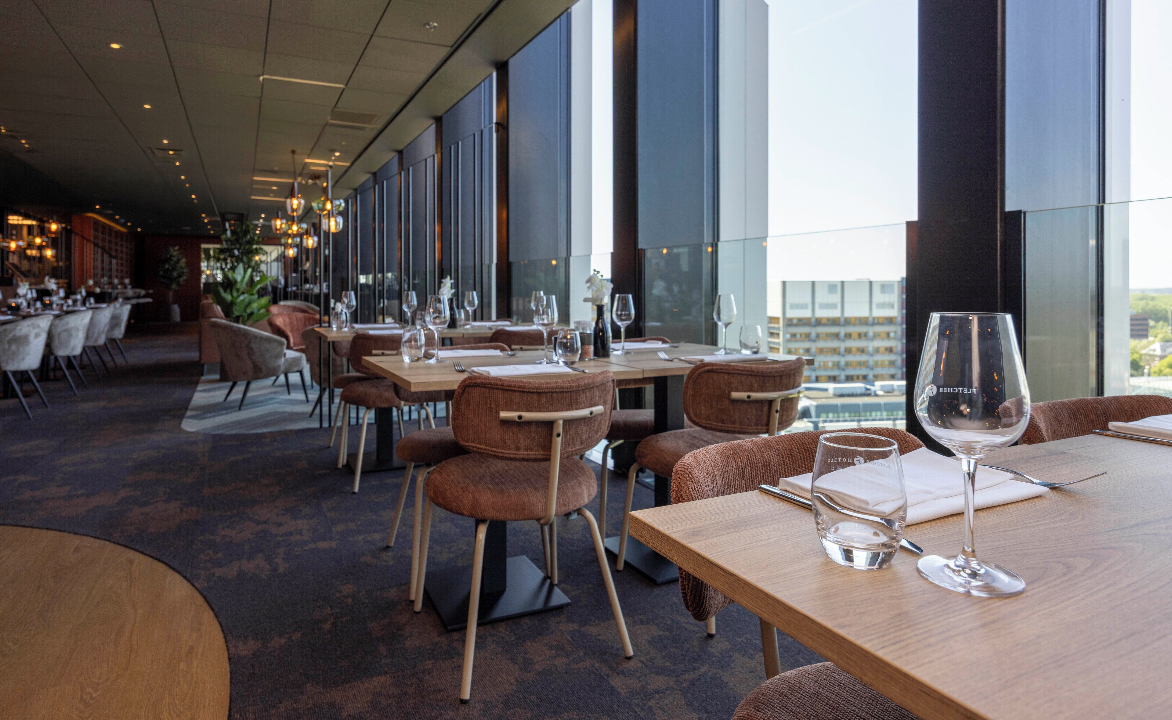 Een modern restaurant met houten tafels en stoelen, grote ramen met uitzicht op de stad, en een bar in de achtergrond.
