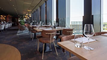 Een modern restaurant met houten tafels en stoelen, grote ramen met uitzicht op de stad, en een bar in de achtergrond.