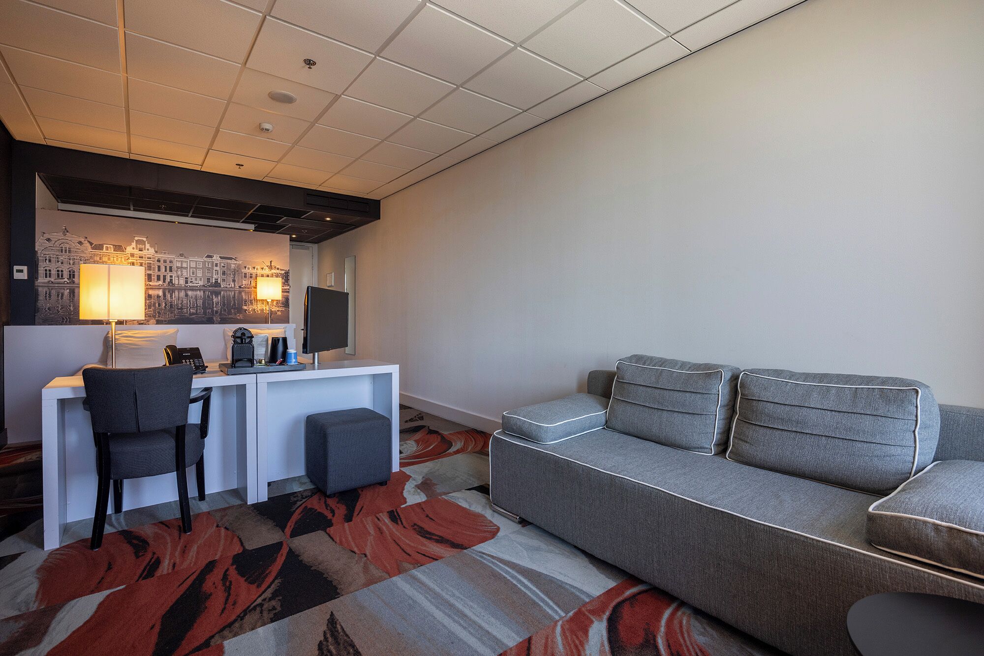 A hotel room with a desk, a chair, a sofa, a patterned rug, and a cityscape artwork on the wall.