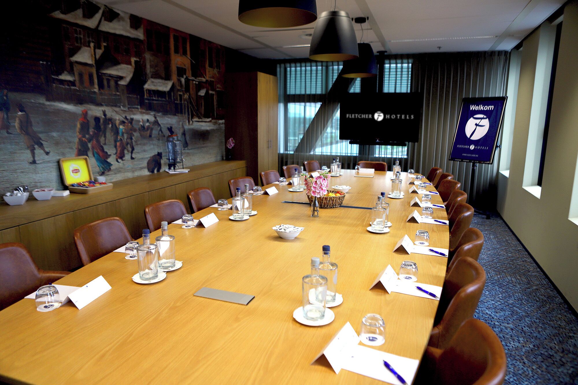 A conference room with a long table, chairs, name cards, water glasses, and a painting on the wall.