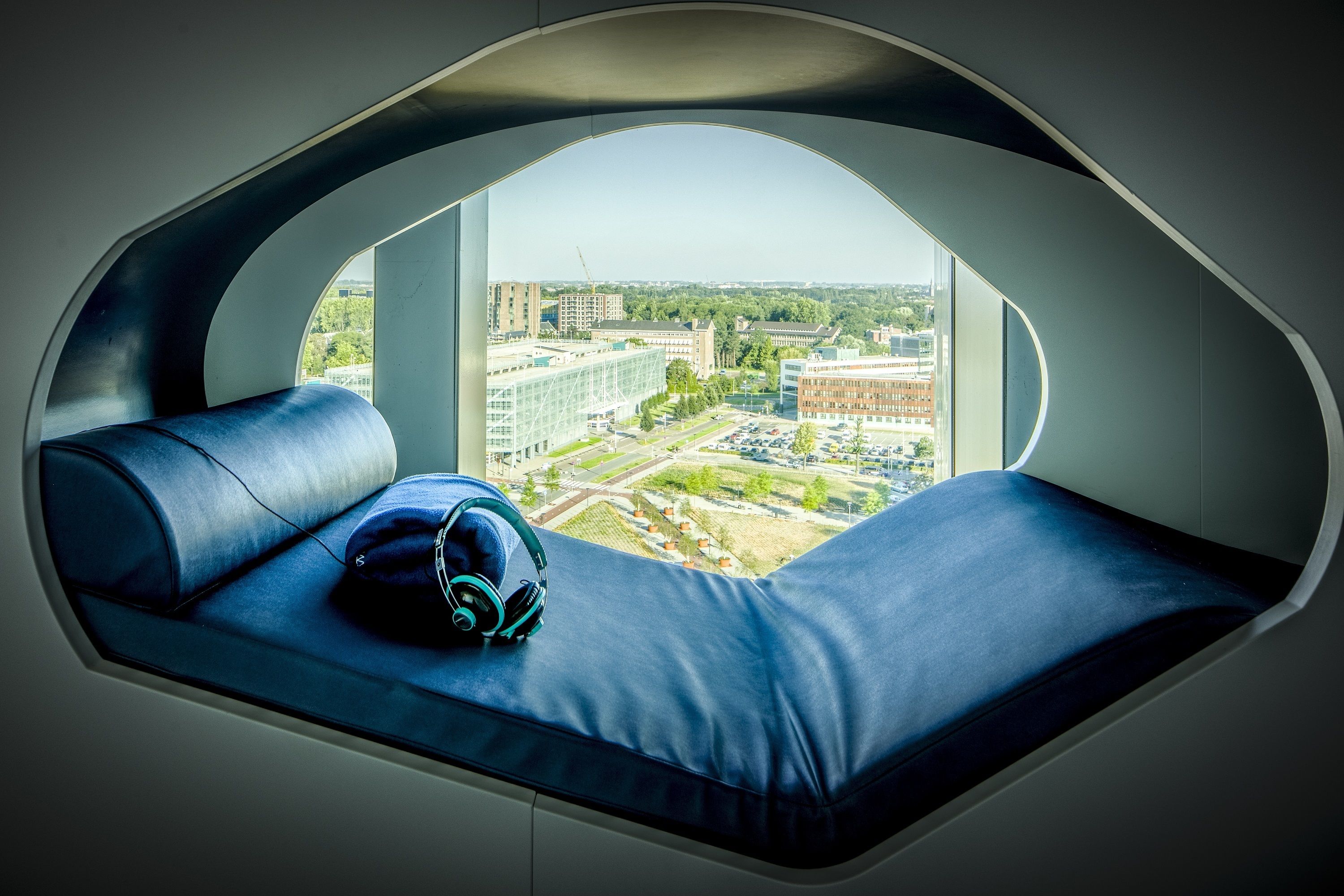 A modern room with a curved window seat, a blue cushioned bench, and a pair of headphones on the bench.