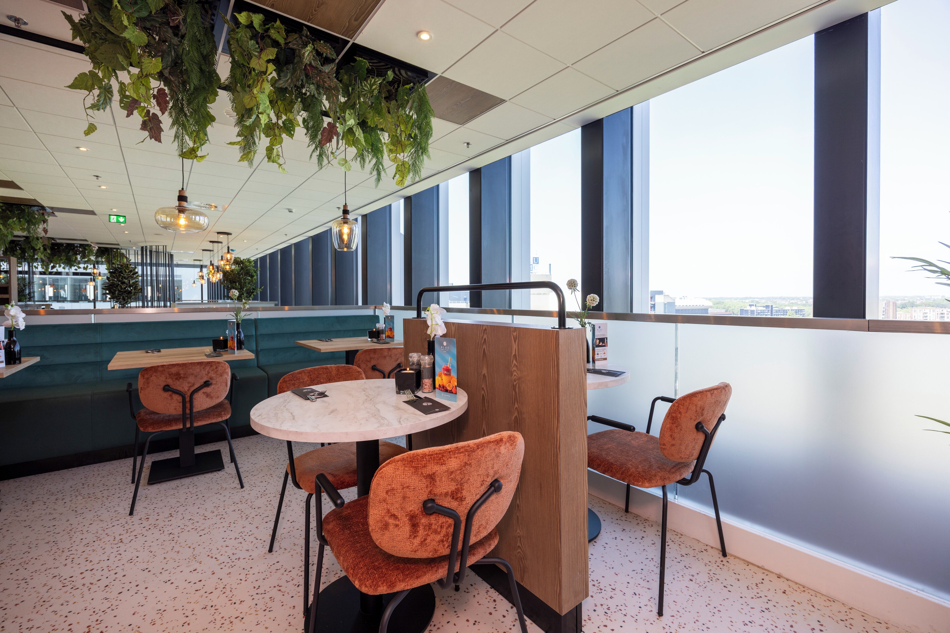 Een modern restaurant met ronde tafels en oranje stoelen, grote ramen met uitzicht over de stad en hangplanten.