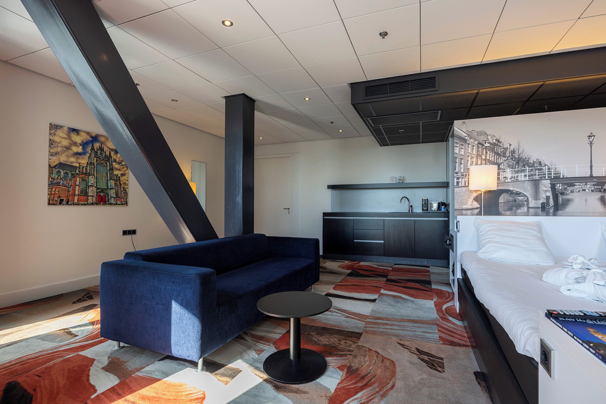 Une chambre d’hôtel moderne comprenant un canapé, une petite table ronde, un lit, un coin cuisine et une fresque murale représentant un paysage urbain.