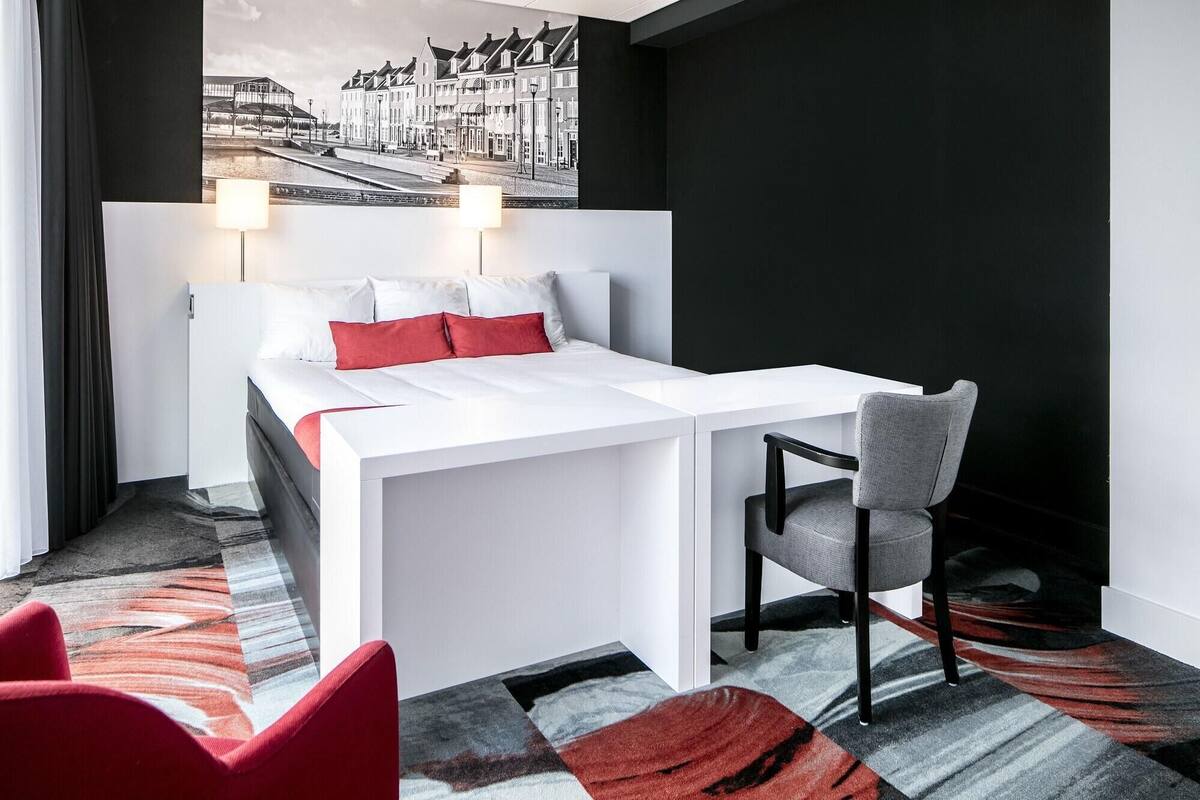 A modern hotel room with a bed, a desk, and a chair. The room has a large window with a view of a street and buildings.