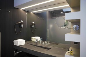 A modern bathroom with a dark wood panel wall, a large mirror, a sink with a faucet, and a towel stack. - Fletcher Wellness - Hotel Helmond (Helmond)
