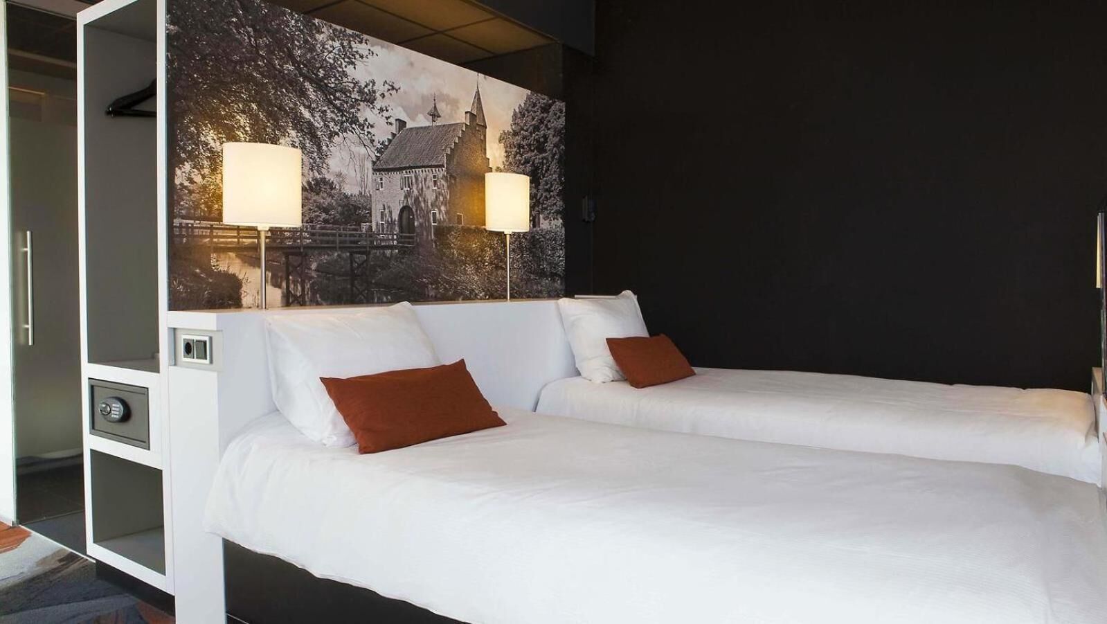 Una cama doble con ropa de cama blanca y dos almohadas marrones, una lámpara de pared y un cuadro grande con un edificio.