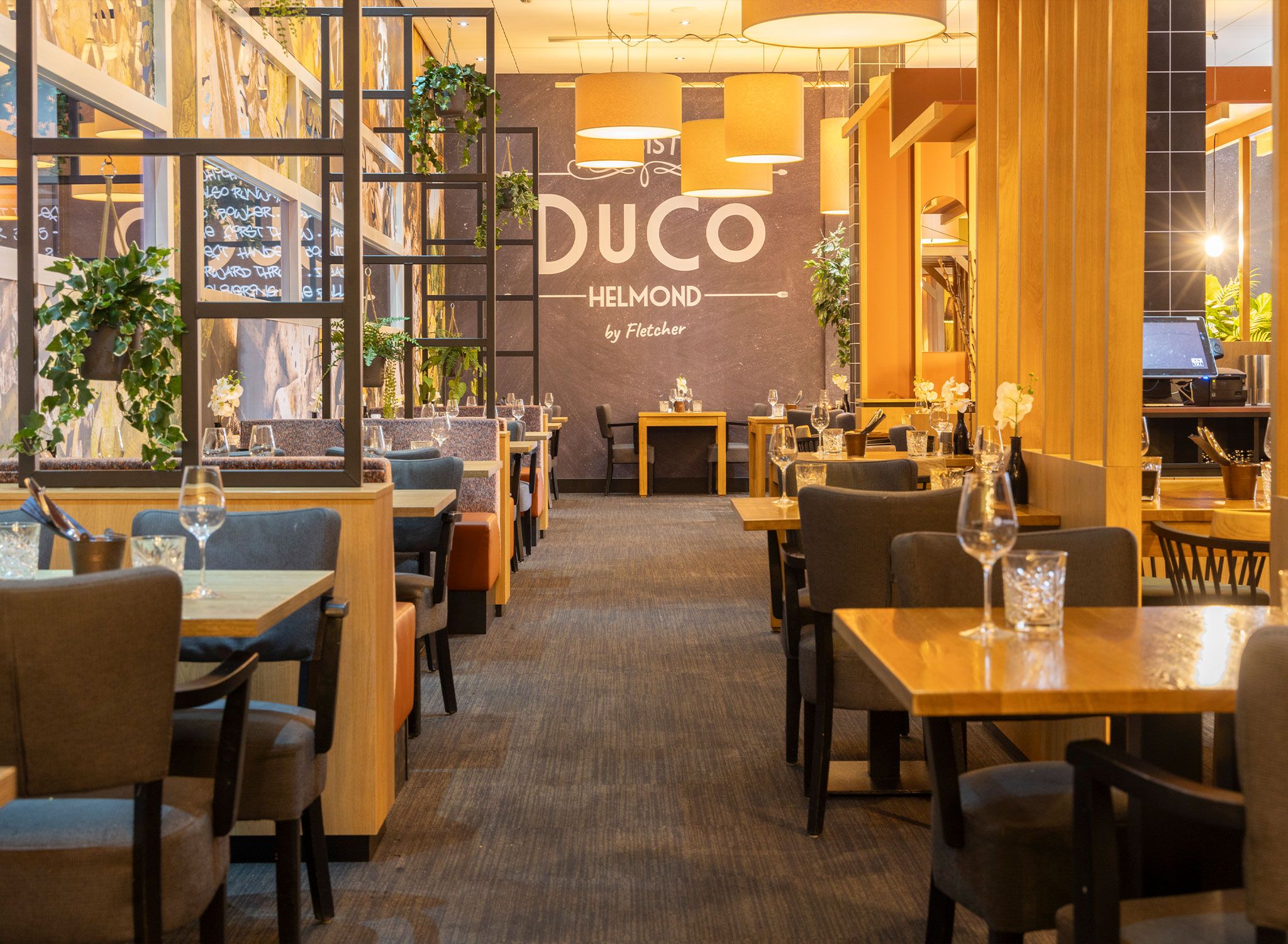 Interno di un ristorante con diversi tavoli e sedie, una parete ampia con il nome 'DUCO HELMOND by Fletcher' e piante pensili.